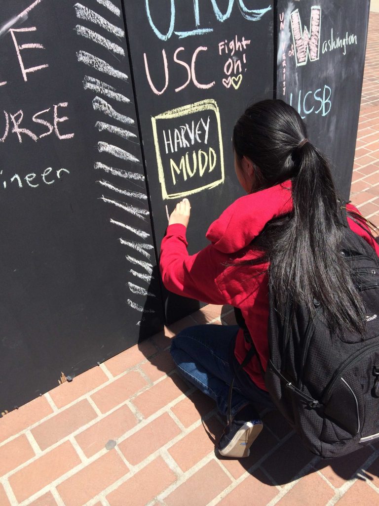 Senior Jennifer Chen writes “UMICH” on a chalkboard in blue and red to show that she will be attending the University of Michigan. Students wrote their colleges on a blackboard to help demonstrate the scope of MVHS.
